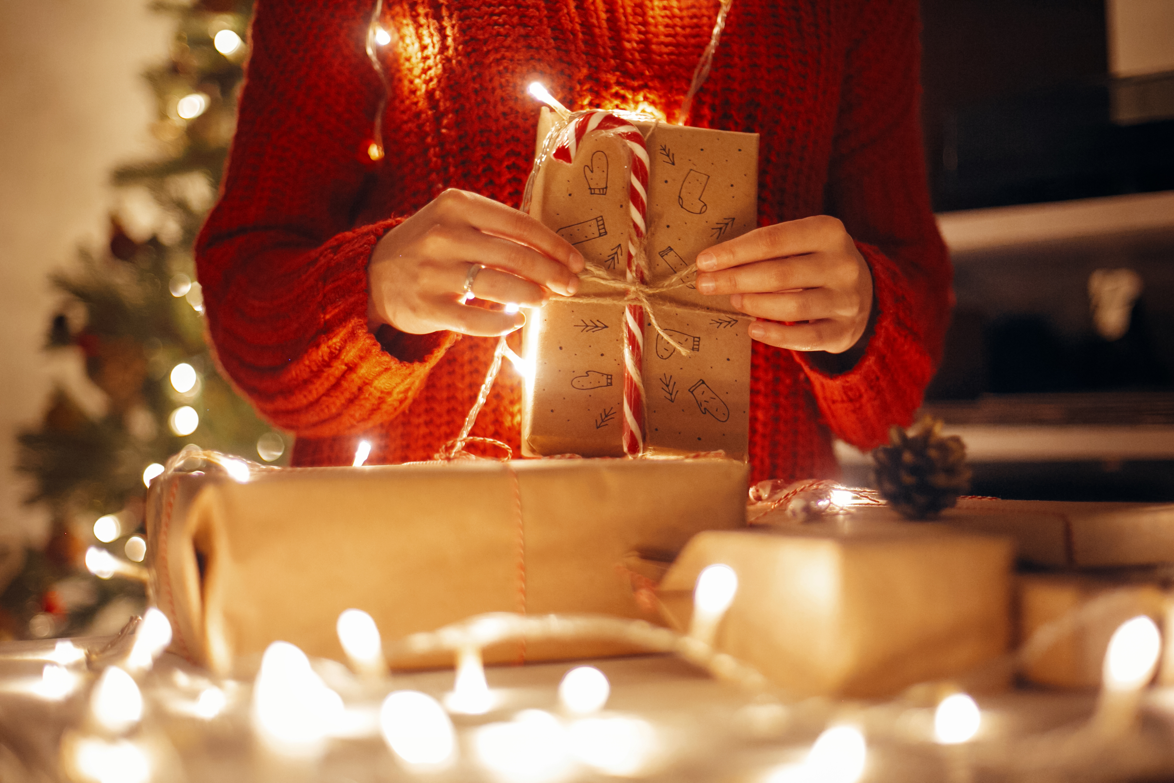 christmas magic moments. hands holding christmas present in lights in evening festive room under tree in illumination. space for text. girl opening modern craft gifts with candy cane