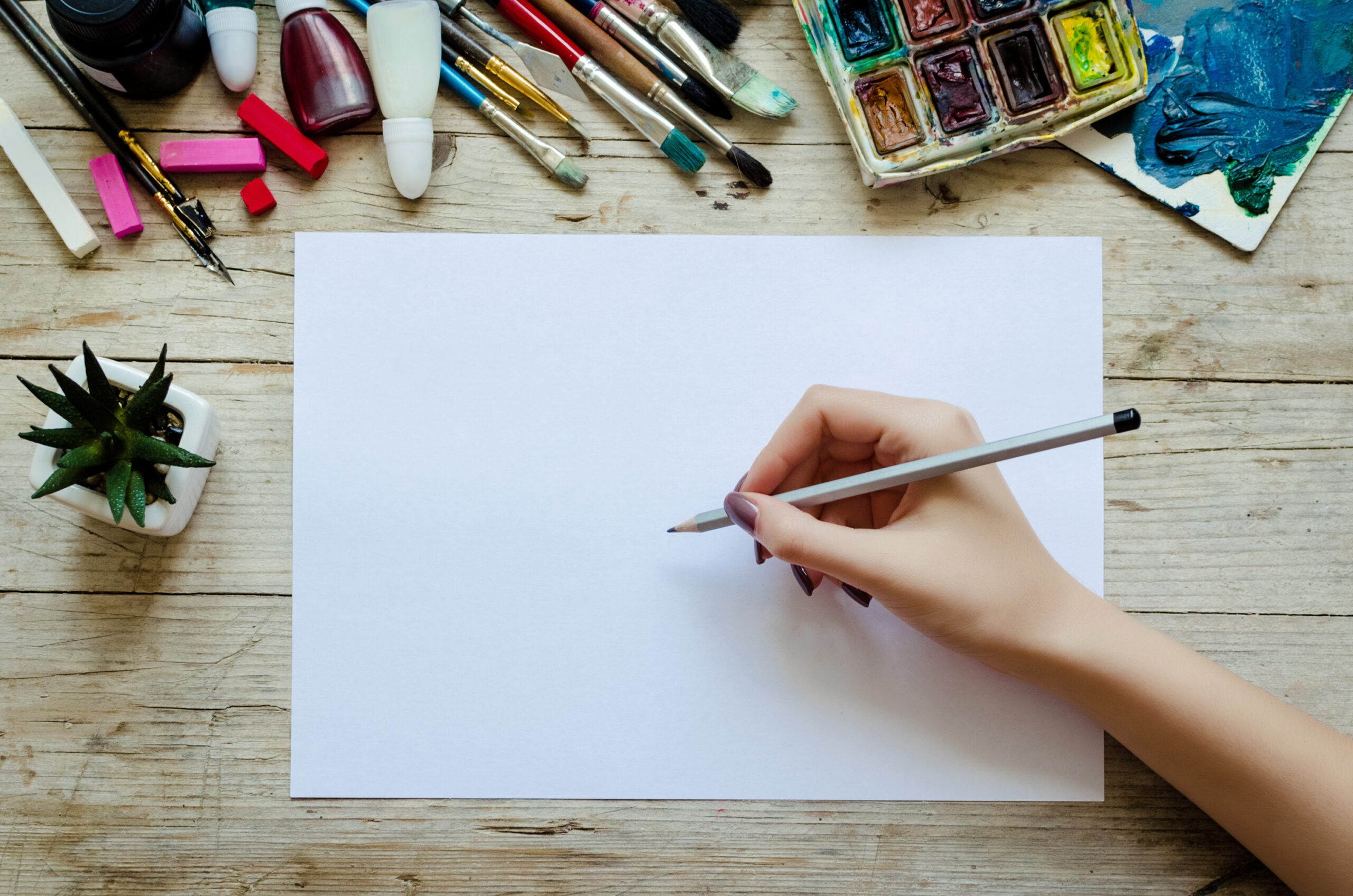 Creative artist workplace flat lay mockup. Top view on table with blank sheet of paper and hand of artist with pencil ready to start sketching or lettering. Art, workshop, drawing concept. Copy space.